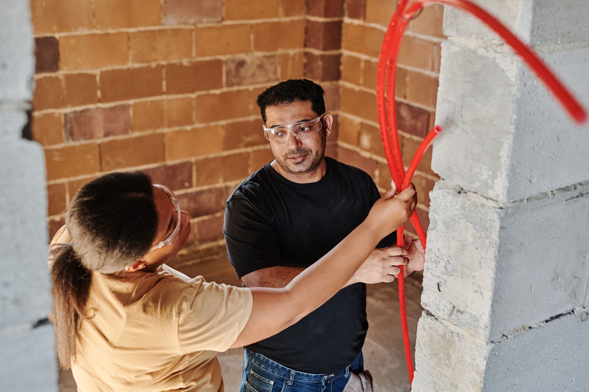 Two workers guiding red tubing through a wall opening